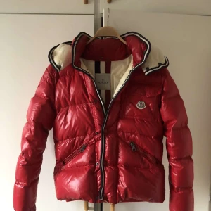 Moncler Branson - Säljer en snygg röd dunjacka från Moncler i mycket bra skick. Jackan har en quiltad design med dragkedja och en mysig huva. Perfekt för kalla vinterdagar och håller dig varm och stilren. Passar perfekt för både vardag och fest! 