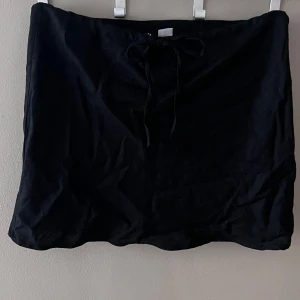 Svart kjol från H&M - Snygg svart kortkjol från H&M i nytt skick. Kjolen har en enkel design med knytband i midjan för en justerbar passform. Perfekt för både vardag och festliga tillfällen. Passar bra till olika toppar och accessoarer för en stilren look.