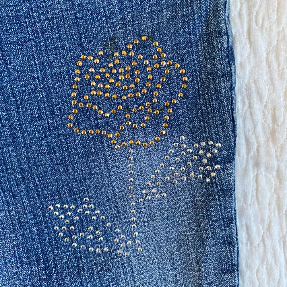 Säljer ett par snygga vintage blå bootcut jeans med  blommigt rhinestones på framsidan. Unika! Midjemått: ca 40 cm, innerbenslängd: ca 77 cm. Jeans & Byxor.