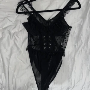 Svart spetsbody från River Island - Snygg svart spetsbody från River Island i storlek 10. Den har ett prickigt mönster och dekorativ snörning framtill. Axelbanden är smala och bodyn har volangdetaljer vid ärmarna. Perfekt för en utekväll eller fest! 🖤