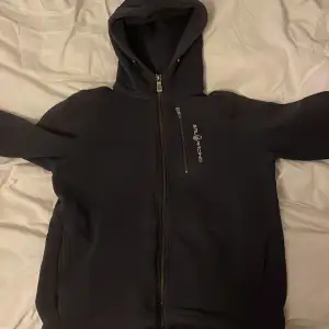 Säljer en mörkblå hoodie från Sail Racing i bra skick det är bara ett litet hål inuti tröjan som ej syns. Den har en dragkedja framtill och en praktisk huva. Perfekt för kyliga dagar eller som ett extra lager. Passar både till vardags och för avslappnade aktiviteter.