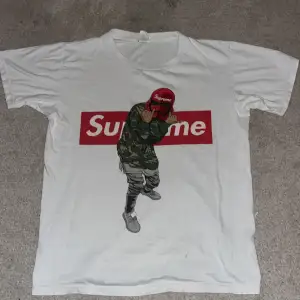 Säljer en vit Supreme t-shirt i bomull med ett coolt tryck av en person i camo och röd keps. Perfekt för streetwear-stilen! T-shirten är kortärmad och har en normal passform. Grym för både vardag och fest.