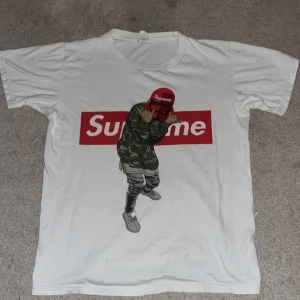 Vit Supreme t-shirt  - Säljer en vit Supreme t-shirt i bomull med ett coolt tryck av en person i camo och röd keps. Perfekt för streetwear-stilen! T-shirten är kortärmad och har en normal passform. Grym för både vardag och fest.