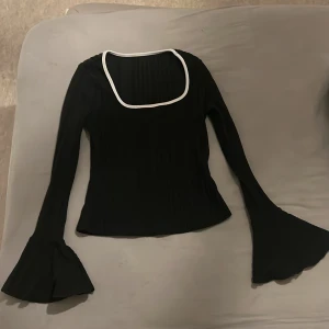 Svart ribbad topp med volangärmar - Snygg svart ribbad topp med långa ärmar och volangdetaljer vid ärmsluten. Den har en unik fyrkantig halsringning med vit kant som ger en cool kontrast. Perfekt för både vardag och fest! 🖤