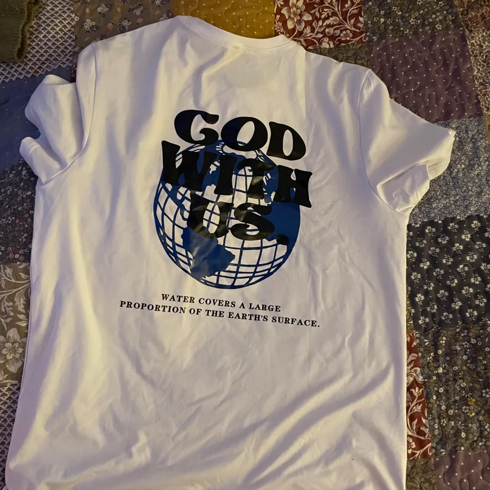 Säljer en vit t-shirt med texten 'GOD WITH US.' tryckt på både fram- och baksidan. På baksidan finns även en jordglob i blått och texten 'WATER COVERS A LARGE PROPORTION OF THE EARTH'S SURFACE.' T-shirten är kortärmad och i bomull, den är oversized . T-shirts.