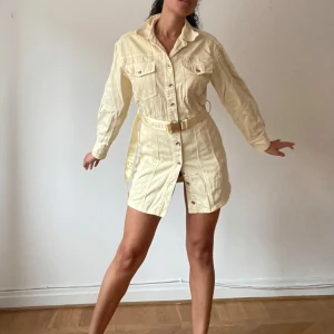 Beige skjortklänning med bälte - Snygg beige skjortklänning i jeansmaterial med långa ärmar och ett matchande bälte i midjan. Perfekt för både vardag och festliga tillfällen. Klänningen har knappar framtill och två bröstfickor som ger en cool och avslappnad look.