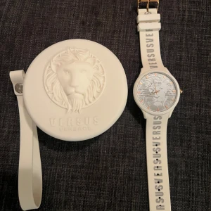 Vit klocka från Versus Versace - Snygg vit klocka från Versus Versace med ett unikt lejonmönster på urtavlan. Armbandet är i vitt med texten 'VERSUS' tryckt längs hela bandet. Klockan kommer i ett matchande runt fodral med lejonmotiv. Perfekt accessoar för att lyfta din stil!
