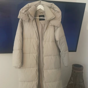 Vinterjacka tjock dunjacka  - Intresse koll på denna tjocka varma dunjackan i fin ljus beige färg, går att knäppa upp på sidorna för att få slitsar på jackan, luvan och kragen går att knäppa så att ingen rusk eller kyla tränger in vid dåligt snöväder, tål - 30 grader kyla 