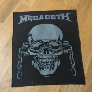 Svart Megadeth tygmärke - Coolt svart tygmärke med Megadeths logga och maskot. Perfekt för att sy på en jacka eller väska för en rockig look. Märket är i bra skick och redo att användas för att ge dina kläder en unik stil.