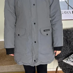 Grå parkajacka från Canada Goose - Säljer en grå parkajacka från Canada Goose i mycket bra skick. Jackan är perfekt för kalla vinterdagar med sin långa design och varma foder. Den har långa ärmar och praktiska fickor framtill. Stängs med både dragkedja och knappar för extra skydd mot kyla. Perfekt för vintern! ❄️Jag som använt jackan är 177 cm lång och har ganska rak kroppsform. Passform kan justeras i midjan till mer smickrande. 
