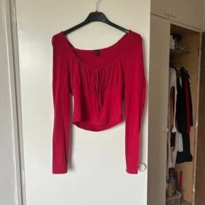 Röd långärmad topp - Säljer en snygg röd långärmad topp i mycket bra skick. Den har en söt knytdetalj framtill och en lätt croppad passform. Perfekt för höst och vår, och funkar både till vardags och fest. Materialet känns mjukt och bekvämt mot huden.