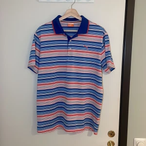 Randig pikétröja från Puma - Säljer en snygg randig pikétröja från Puma i blått, rött och vitt. Perfekt för sommardagar eller en avslappnad dag på stan. Tröjan har en klassisk krage och knappar framtill. Den är i mycket bra skick och redo för nya äventyr! Pris går alltid att förhandlas !