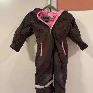 Brun och rosa overall från HM stl 110 - Säljer en brun/rosa  overall från  HM med rosa foder och detaljer. Perfekt för kalla dagar med sin varma och skyddande design. Overallen har en dragkedja framtill och flera praktiska fickor. Den är i bra skick och redo för nya äventyr!