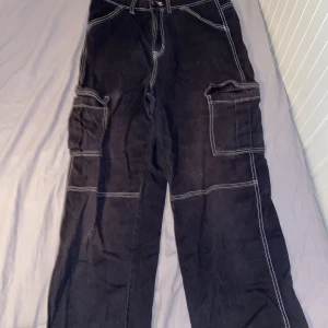Svarta cargopants från H&M Divided - Säljer ett par svarta cargopants från H&M Divided i storlek 38. Byxorna har vita kontrastsömmar och praktiska fickor på sidorna. De är i bra skick men har ett litet hål på båda benen. Priset går att diskutera.
