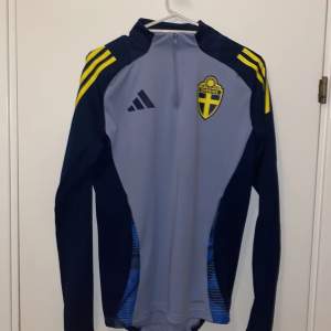 Säljer en snygg grå och marinblå träningströja från Adidas med Sveriges landslagsemblem på bröstet. Tröjan har en dragkedja vid halsen och gula detaljer på axlarna. Perfekt för fotbollsträning eller andra sportaktiviteter. Slim fit för en skön passform! (Använd ett fåtal gånger 1-7 ggr köptes i somras)