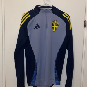 Sverige träningströja Grå och marinblå träningströja från Adidas  - Säljer en snygg grå och marinblå träningströja från Adidas med Sveriges landslagsemblem på bröstet. Tröjan har en dragkedja vid halsen och gula detaljer på axlarna. Perfekt för fotbollsträning eller andra sportaktiviteter. Slim fit för en skön passform! Går för 750kr ny (Använd ett fåtal gånger 1-7 ggr köptes i somras)