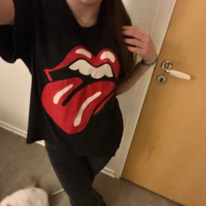 Svart t-shirt med rött tryck - Cool svart t-shirt med det ikoniska röda tungan-trycket. Perfekt för en avslappnad stil eller konsertkväll. T-shirten har en normal passform och är kortärmad. Ett måste för alla rockälskare!