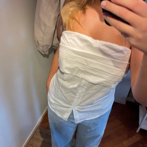 Vit axelbandslös topp - Säljer en stilren vit axelbandslös topp. Den har en enkel och elegant design med en knäppning framtill. Perfekt för sommardagar eller en avslappnad festkväll. Toppen är oanvänd och passar perfekt till jeans eller kjol.