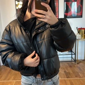 Svart pufferjacka Zara  - Säljer en snygg svart pufferjacka i syntetmaterial. Jackan har en quiltad design med dragkedja och knappar framtill. Den är perfekt för höst och vinter, och har långa ärmar med elastiska muddar för extra värme. Passformen är normal och den är superbekväm att ha på sig. Perfekt för att hålla stilen när det är kallt ute!