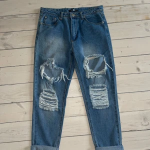 Blå jeans med slitningar - Säljer ett par blå jeans med coola slitningar på framsidan. De har en avslappnad passform och är perfekt för en casual look. Jeansen är uppvikta vid bensluten för en extra touch av stil, små i storleken