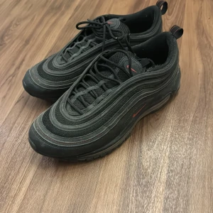Svarta Nike Air Max 97 med röda detaljer - Säljer ett par svarta Nike Air Max 97 med snygga röda detaljer. Skorna har en ikonisk design med synlig Air-enhet i sulan för extra komfort. De är i bra skick med lite slitage på sulan. Perfekta för både vardag och träning!