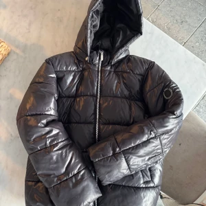 Svart pufferjacka från ONLY - Säljer en snygg svart pufferjacka från ONLY. Den har en glansig finish och en praktisk huva. Jackan är perfekt för kyliga dagar och har en dragkedja framtill. Den är i bra skick och redo för nya äventyr!