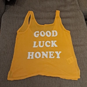Gul linne med text - Säljer en cool gul linne med texten 'GOOD LUCK HONEY' i vitt på framsidan. Perfekt för sommardagar eller en avslappnad look. Linnet är ärmlöst och har en lös passform.