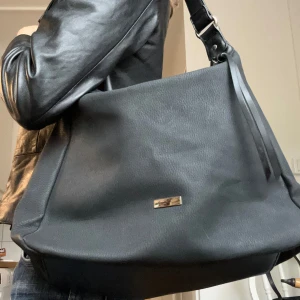 Svart faux skin handväska  - Svart faux skin handväska i nyskick och ej använd! Har tofs på dragkedjan, en axelrem och mycket utrymme med ficka innanför. Har ingen label. 