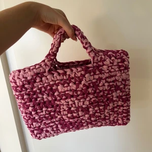 Handgjord rosa stickad väska - Säljer en superfin handgjord virkad väska. Den är i olika nyanser av rosa och har ett unikt flätat mönster. Perfekt för sommarens utflykter eller som en söt accessoar till vardagsoutfiten. Väskan har två handtag 