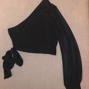 Svart oneshoulder topp med knyt - Säljer en snygg svart oneshoulder topp med knytning på sidan. Den har en lång ärm i ett transparent material. Köpt på shein, använd ett fåtal gånger så i väldigt fint skick! 