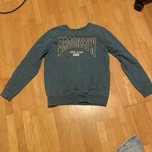 Blå sweatshirt från Kappahl - Säljer en blå sweatshirt från Kappahl med texten 'Brooklyn New York 1978' på framsidan. Tröjan har långa ärmar och en rund halsringning. Perfekt för en avslappnad stil.