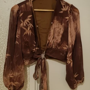 Brun blus med mönster  - Brun kort blus med mönster av blad i ljusare nyans. Fin att ha över en klänning eller tröja. Nedre delen kan knytas runt magen och den går även att knäppa.