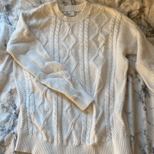 Vit stickad tröja från Hollister - Säljer en snygg vit stickad tröja från Hollister med kabelstickat mönster. Tröjan har långa ärmar och ribbade muddar vid ärmslut och nederkant. Perfekt för kyligare dagar! Sälja på grund av inte till användning 