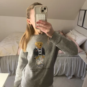 Sweatshirt från Ralph Lauren - Grå sweatshirt från Ralph Lauren med Polo Bear-motivet på framsidan. Tröjan har långa ärmar och en rund halsringning. Tröjan är använd men inga defekter. 