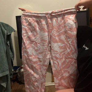 Rosa och vita mjukisbyxor med mönster - Säljer ett par bekväma mjukisbyxor i rosa och vitt med ett snyggt abstrakt mönster. Byxorna har en elastisk midja med snörning för extra komfort. Perfekta för en avslappnad dag hemma.