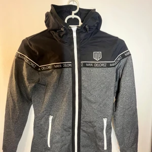 Grå och svart jacka från Maya Delorez - Snygg grå och svart tröja från Maya Delorez med dragkedja och huva. Jackan har vita detaljer och logotyp på. Använd två gånger. 