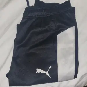 Snygga svarta träningsbyxor från Puma med vita detaljer längs sidorna. Byxorna har en elastisk midja med snörning för bästa passform och det ikoniska Puma-loggan på benet. Perfekta för träning eller en avslappnad dag.