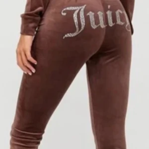 Bruna velourbyxor från Juicy Couture - Snygga bruna velourbyxor från Juicy Couture med glittrig text på baksidan. Byxorna har en tight passform och är perfekta för en avslappnad stil. De är mjuka och bekväma, vilket gör dem till ett måste i garderoben.
