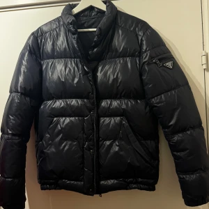 Mörkblå pufferjacka från Prada - Nypris ca: 16000kr Säljer en mörkplå pufferjacka från Prada med dragkedja och tryckknappar. Jackan har en klassisk design med långärmade ärmar och en hög krage. Den är tillverkad i ett glansigt nylon och har Pradas logotyp på ärmen. Perfekt för kyliga dagar.