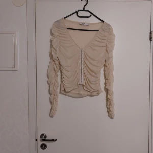 Beige blus med rynkdetaljer - Säljer en elegant beige blus från NA-KD med rynkdetaljer och v-ringning. Blusen har långa ärmar och knäppning framtill. Perfekt för en stilren look.