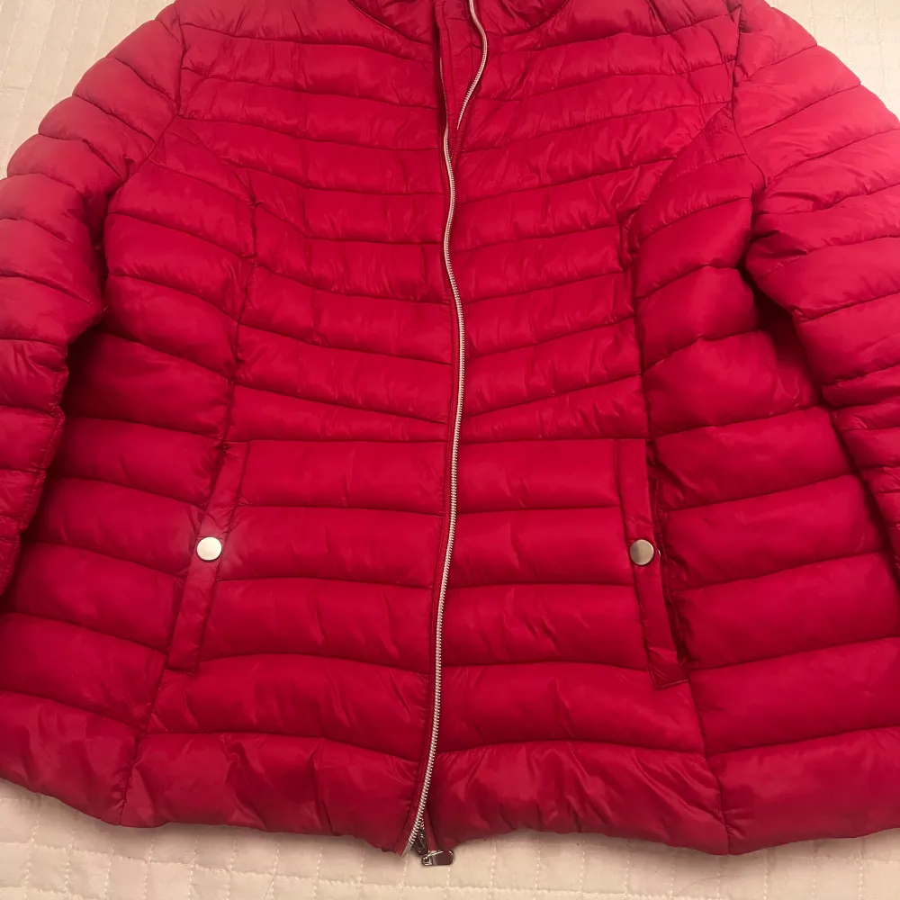 Snygg röd dunjacka med dragkedja framtill och två fickor med knappar. Perfekt för kyliga dagar och ger en skön värme. Jackan har en klassisk design med quiltade detaljer och långärmade ärmar.. Takit.