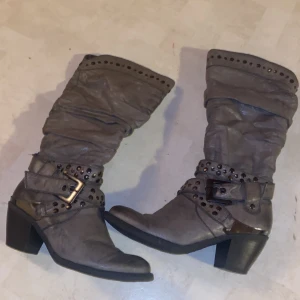 Grå boots med nitar och bälte - Snygga grå boots med dekorativa nitar och bälte. De har en robust klack och en rynkad design som ger en cool look. Perfekta för att ge din outfit en edgy touch.