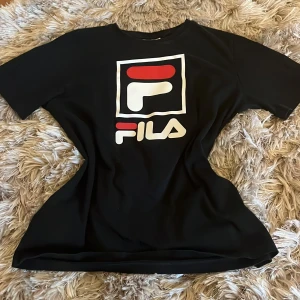 Svart t-shirt från FILA - Säljer en svart t-shirt från FILA med deras ikoniska logga i vitt och rött på framsidan. T-shirten har en klassisk passform och korta ärmar, perfekt för en avslappnad men lyxig stil. Har använt 2 gånger och den tvättas och stryks innan den skickad iväg! Köpte för 300kr men säljer för 150!! Priset kan diskuteras💗💗Den passar för personer som har xs, s och m