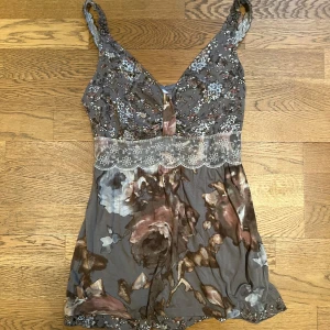 Blommig linne i grå och rosa toner - Säljer ett vackert linne med blommigt mönster i grå och rosa toner. Linnet har spetsdetaljer under bysten och breda axelband. Perfekt för en somrig look!