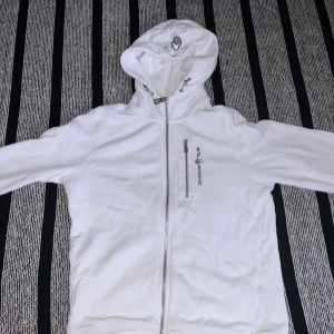 Vit huvtröja från Sail Racing - Säljer en stilren vit huvtröja från Sail Racing. Tröjan har en dragkedja framtill och en justerbar huva. Perfekt för en avslappnad stil. Den har långa ärmar och en liten logotyp på bröstet.