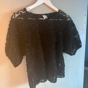 Svart blus från Visual Clothing Project - Säljer en elegant svart blus från Visual Clothing Project. Blusen har ett unikt blommigt mönster och är tillverkad i ett transparent material. Den har trekvartsärmar och en lös passform, perfekt för en stilfull look. Säljer pågrund av flytt och för mycket kläder 😁😁😁