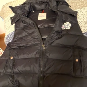 Svart dunväst från Moncler - Snygg svart dunväst från Moncler med dragkedja och tryckknappar. Västen har en quiltad design och en avtagbar huva. Perfekt för kyligare dagar.                                     Skriv för info eller vid intresse!