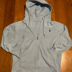 Ljusblå hoodie från Polo Ralph Lauren - Säljer en stilren ljusblå hoodie från Polo Ralph Lauren med dragkedja och broderad logga på bröstet. Hoodien har en justerbar huva med snören och praktiska fickor framtill. Perfekt för en avslappnad stil.