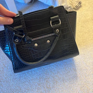 Svart handväska med krokodilmönster - Snygg svart handväska med krokodilmönster och dragkedja. Väskan har två handtag och en stilren design som passar perfekt för en chic look. Perfekt för att bära dina dagliga nödvändigheter med stil. Kommer ej ihåg vart jag köpte det då jag har haft den i cirka 3 år, typ aldrig använt då jag har så mycket väskor. Säljer pågrund av flytt och för mycket kläder 😁