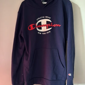 Mörkblå hoodie från Champion - Säljer en snygg mörkblå hoodie från Champion med deras ikoniska logga i rött och vitt på bröstet. Hoodien har en klassisk känguruficka och långa ärmar. Perfekt för en avslappnad stil.